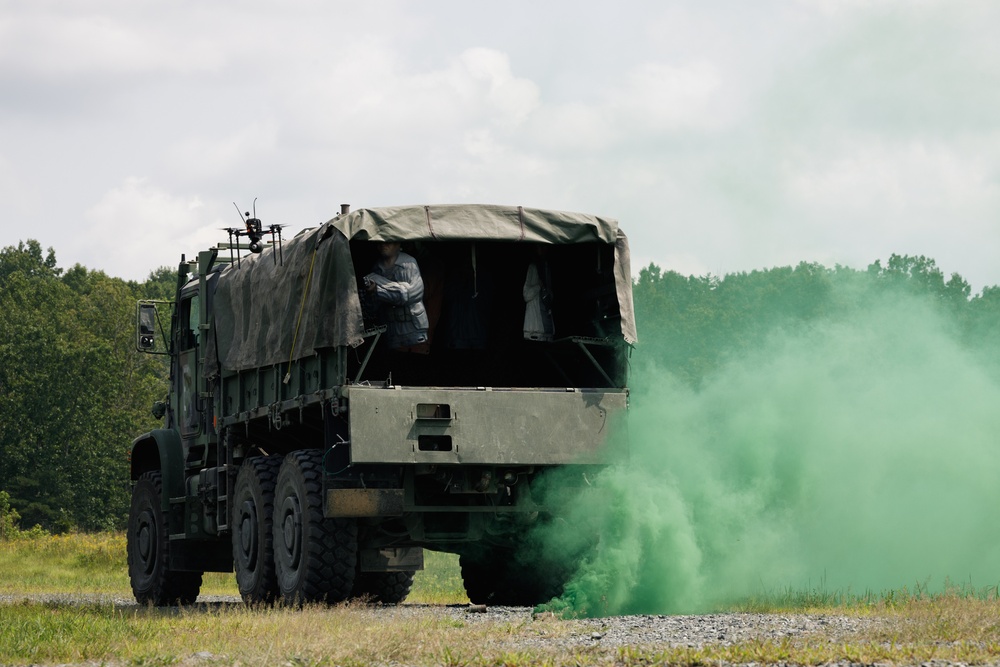 A military cargo truck, consistent with a LMTV or FMTV-type vehicle, is positioned on a gravel surface with bright green smoke emanating from beneath or beside the rear of the vehicle. A person in what appears to be a protective or military uniform is visible in the rear cargo area under a canvas tarp cover. The scene appears to be a military training exercise, with the green smoke likely from a smoke grenade used for signaling or simulation purposes.
