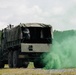 A military cargo truck, consistent with a LMTV or FMTV-type vehicle, is positioned on a gravel surface with bright green smoke emanating from beneath or beside the rear of the vehicle. A person in what appears to be a protective or military uniform is visible in the rear cargo area under a canvas tarp cover. The scene appears to be a military training exercise, with the green smoke likely from a smoke grenade used for signaling or simulation purposes.