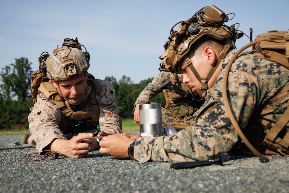 Two military personnel in full combat gear are lying prone on a paved surface, closely examining a cylindrical metallic object placed between them. Both are wearing MARPAT (Marine pattern) camouflage uniforms with tactical helmets equipped with night vision mounts and communication devices. The setting appears to be an outdoor training environment with trees and open sky visible in the background.