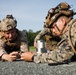 Two military personnel in full combat gear are lying prone on a paved surface, closely examining a cylindrical metallic object placed between them. Both are wearing MARPAT (Marine pattern) camouflage uniforms with tactical helmets equipped with night vision mounts and communication devices. The setting appears to be an outdoor training environment with trees and open sky visible in the background.