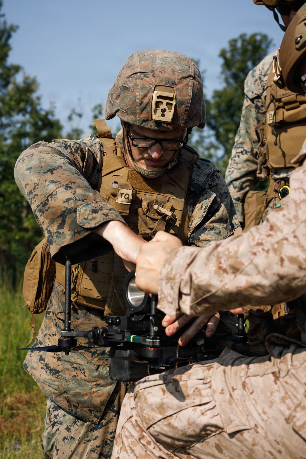 Two or more military personnel in MARPAT (Marine pattern) camouflage uniforms are handling a small drone or unmanned aerial system (UAS). The central figure wears a combat helmet with a NVG mount, ballistic eyewear, and a plate carrier vest marked 'TQ'. They appear to be assembling, inspecting, or preparing the drone for operation in an outdoor field environment.