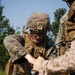 Two or more military personnel in MARPAT (Marine pattern) camouflage uniforms are handling a small drone or unmanned aerial system (UAS). The central figure wears a combat helmet with a NVG mount, ballistic eyewear, and a plate carrier vest marked 'TQ'. They appear to be assembling, inspecting, or preparing the drone for operation in an outdoor field environment.