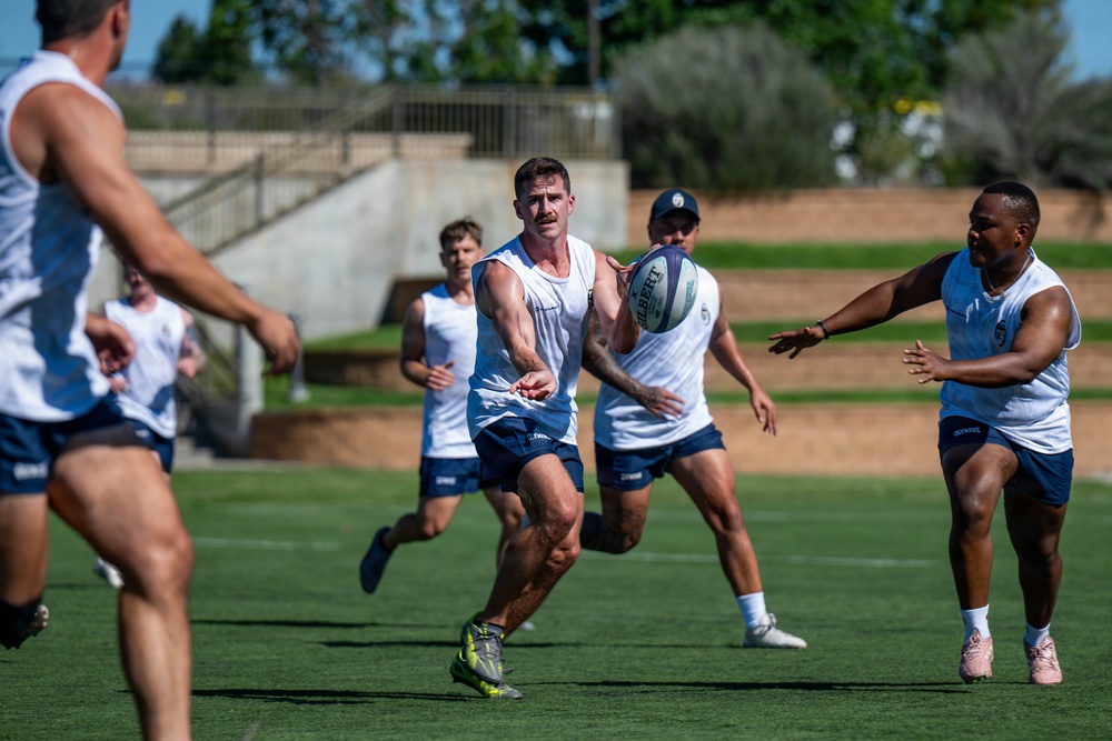 A group of athletes are engaged in a rugby training session on a well-maintained grass field under bright sunlight. The central player is in the act of passing a Gilbert rugby ball laterally to a teammate on the right. All players are wearing matching white sleeveless tops and dark blue shorts, indicating a team practice environment.