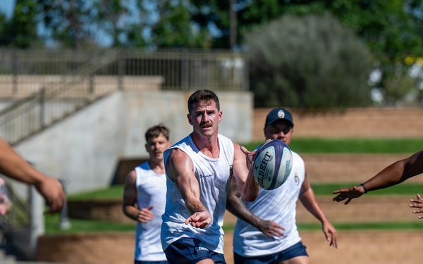 A group of athletes are engaged in a rugby training session on a well-maintained grass field under bright sunlight. The central player is in the act of passing a Gilbert rugby ball laterally to a teammate on the right. All players are wearing matching white sleeveless tops and dark blue shorts, indicating a team practice environment.