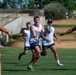 A group of athletes are engaged in a rugby training session on a well-maintained grass field under bright sunlight. The central player is in the act of passing a Gilbert rugby ball laterally to a teammate on the right. All players are wearing matching white sleeveless tops and dark blue shorts, indicating a team practice environment.