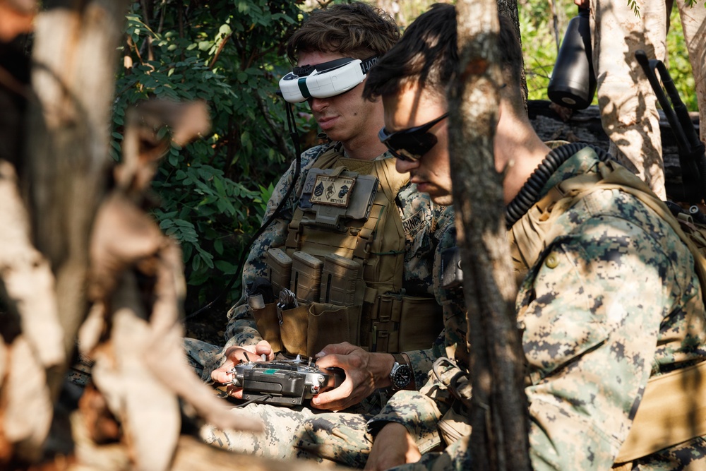 Two military personnel in digital camouflage uniforms are operating what appears to be an FPV (First-Person View) drone system in a wooded outdoor environment. The soldier in the foreground is wearing FPV goggles on his forehead and holding a drone remote controller, while a second soldier wearing sunglasses observes nearby. Both are equipped with tactical vests and gear consistent with U.S. Marine Corps uniforms.