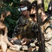 Two military personnel in digital camouflage uniforms are operating what appears to be an FPV (First-Person View) drone system in a wooded outdoor environment. The soldier in the foreground is wearing FPV goggles on his forehead and holding a drone remote controller, while a second soldier wearing sunglasses observes nearby. Both are equipped with tactical vests and gear consistent with U.S. Marine Corps uniforms.