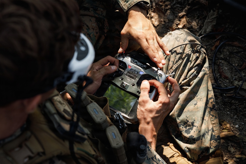 Two military personnel in camouflage uniforms are handling a Futaba brand radio transmitter/remote control unit, likely used for operating an unmanned aerial vehicle (drone) or similar remote-controlled system. The image is taken from an overhead angle in an outdoor, wooded environment. One individual appears to be instructing or assisting the other with the device.