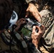 Two military personnel in camouflage uniforms are handling a Futaba brand radio transmitter/remote control unit, likely used for operating an unmanned aerial vehicle (drone) or similar remote-controlled system. The image is taken from an overhead angle in an outdoor, wooded environment. One individual appears to be instructing or assisting the other with the device.