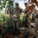 A group of military personnel in camouflage uniforms are gathered in a wooded outdoor setting, appearing to be engaged in a training exercise or field operation. Senior personnel appear to be observing or instructing others who are positioned lower, possibly in a trench or foxhole. One individual is wearing what appears to be VR/AR goggles, suggesting a technology-integrated training scenario.