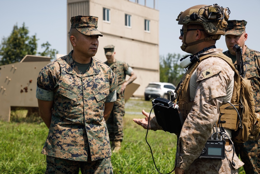 The image shows U.S. Marines in a field training or operational environment, with one Marine in standard utility uniform conversing with a fully equipped Marine wearing tactical gear including a combat helmet with mounted accessories and body armor. A third Marine is visible in the background. The setting appears to be a military training facility with a concrete structure visible in the background.