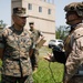 The image shows U.S. Marines in a field training or operational environment, with one Marine in standard utility uniform conversing with a fully equipped Marine wearing tactical gear including a combat helmet with mounted accessories and body armor. A third Marine is visible in the background. The setting appears to be a military training facility with a concrete structure visible in the background.