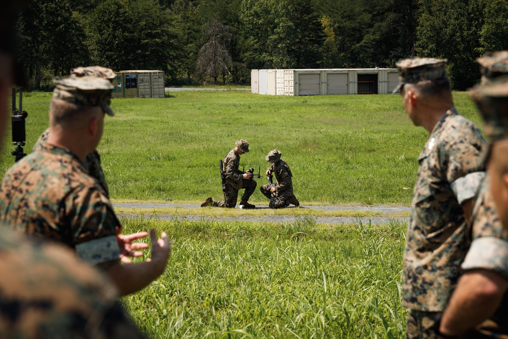 Military personnel in woodland camouflage uniforms are conducting what appears to be a drone or unmanned aerial system (UAS) demonstration or training exercise in an open field. Two soldiers in the middle ground are kneeling and handling a small drone/UAS device, while observers in the foreground watch and discuss the activity. The setting appears to be a military training area with storage containers and tree lines visible in the background.