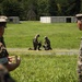 Military personnel in woodland camouflage uniforms are conducting what appears to be a drone or unmanned aerial system (UAS) demonstration or training exercise in an open field. Two soldiers in the middle ground are kneeling and handling a small drone/UAS device, while observers in the foreground watch and discuss the activity. The setting appears to be a military training area with storage containers and tree lines visible in the background.
