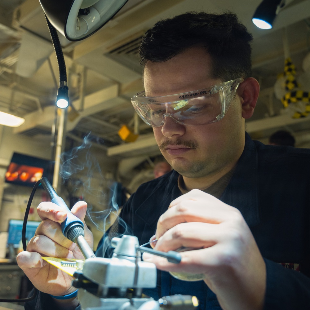 A technician wearing clear safety glasses is performing soldering work on a yellow circuit board in what appears to be an industrial or military workspace. Visible solder smoke rises from the work area as the person carefully manipulates components with precision tools. The environment is well-lit with task lighting and magnification equipment visible overhead.