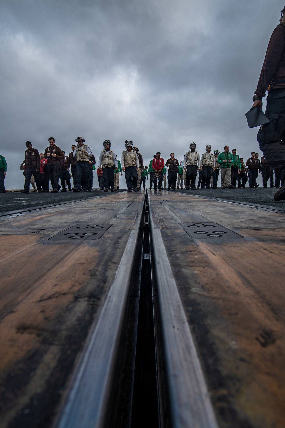 A low-angle ground-level photograph taken on an aircraft carrier flight deck, looking along a catapult launch track slot toward a large group of personnel. The personnel are wearing various colored flight deck jerseys and safety gear, suggesting a multi-role crew assembly. The overcast sky and wet deck surface indicate cloudy maritime weather conditions.