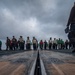 A low-angle ground-level photograph taken on an aircraft carrier flight deck, looking along a catapult launch track slot toward a large group of personnel. The personnel are wearing various colored flight deck jerseys and safety gear, suggesting a multi-role crew assembly. The overcast sky and wet deck surface indicate cloudy maritime weather conditions.