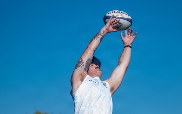 A rugby lineout is being executed during what appears to be a practice or match session on a grass field under clear blue skies. One player is being lifted by teammates to catch a Gilbert rugby ball at its highest point. Players are wearing white sleeveless jerseys and navy blue shorts, with visible tattoos and an 'America 250' logo on one jersey.