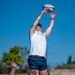 A rugby lineout is being executed during what appears to be a practice or match session on a grass field under clear blue skies. One player is being lifted by teammates to catch a Gilbert rugby ball at its highest point. Players are wearing white sleeveless jerseys and navy blue shorts, with visible tattoos and an 'America 250' logo on one jersey.