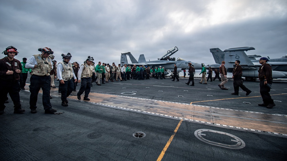 The image shows a large group of naval personnel conducting what appears to be a Foreign Object Debris (FOD) walkdown on the flight deck of a US Navy aircraft carrier. Multiple crew members in various colored jerseys and vests are walking in formation across the deck. At least two F/A-18 Hornet or Super Hornet jet aircraft are visible parked on the deck in the background.