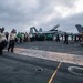 The image shows a large group of naval personnel conducting what appears to be a Foreign Object Debris (FOD) walkdown on the flight deck of a US Navy aircraft carrier. Multiple crew members in various colored jerseys and vests are walking in formation across the deck. At least two F/A-18 Hornet or Super Hornet jet aircraft are visible parked on the deck in the background.