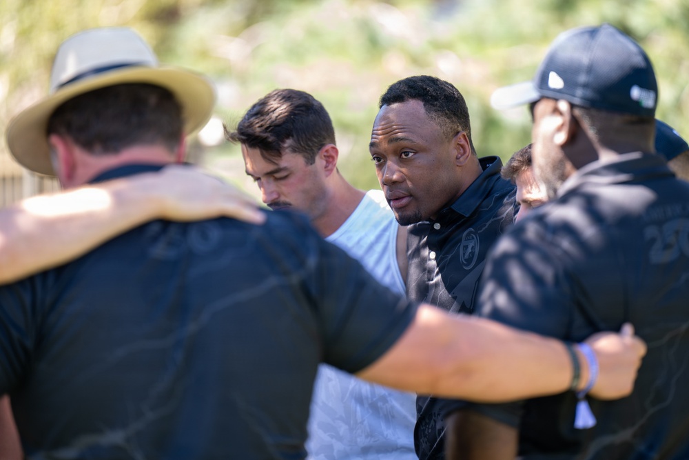 A group of men are gathered in a close huddle outdoors, with arms around each other's shoulders. The central figure, a man in a dark polo shirt, appears to be speaking or leading the group. The setting appears to be an outdoor daytime gathering with green foliage visible in the background.
