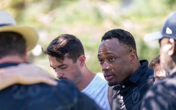 A group of men are gathered in a close huddle outdoors, with arms around each other's shoulders. The central figure, a man in a dark polo shirt, appears to be speaking or leading the group. The setting appears to be an outdoor daytime gathering with green foliage visible in the background.