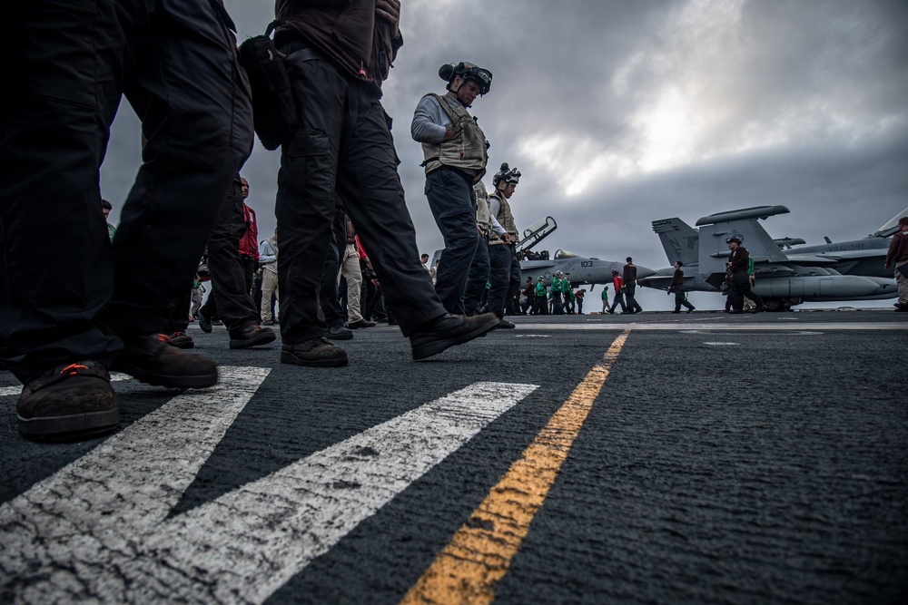 Personnel are walking across the flight deck of a U.S. Navy aircraft carrier under overcast skies. Multiple jet aircraft, including what appears to be an F/A-18 Hornet or Super Hornet marked '103', are parked on the deck. Crew members in various colored jerseys — indicating different roles — are visible in the background.