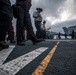 Personnel are walking across the flight deck of a U.S. Navy aircraft carrier under overcast skies. Multiple jet aircraft, including what appears to be an F/A-18 Hornet or Super Hornet marked '103', are parked on the deck. Crew members in various colored jerseys — indicating different roles — are visible in the background.