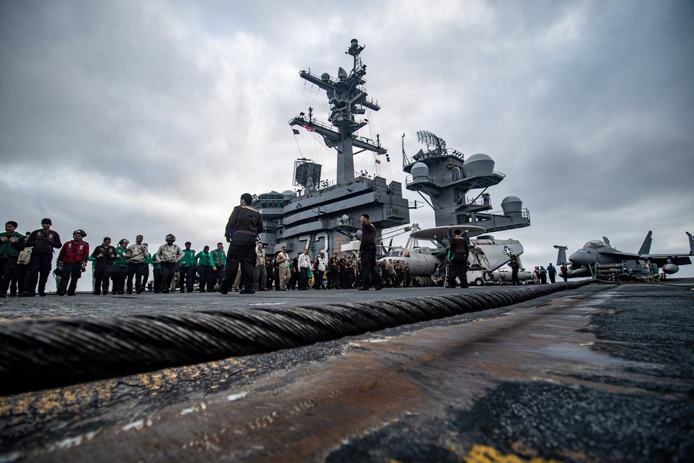 The image shows the flight deck of a US Navy aircraft carrier with a large group of personnel gathered near the island superstructure. A thick arresting wire cable is prominently visible in the foreground, stretching across the deck. At least two aircraft are visible on the deck, including what appears to be an E-2 Hawkeye and an F/A-18 Hornet/Super Hornet.