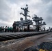 The image shows the flight deck of a US Navy aircraft carrier with a large group of personnel gathered near the island superstructure. A thick arresting wire cable is prominently visible in the foreground, stretching across the deck. At least two aircraft are visible on the deck, including what appears to be an E-2 Hawkeye and an F/A-18 Hornet/Super Hornet.