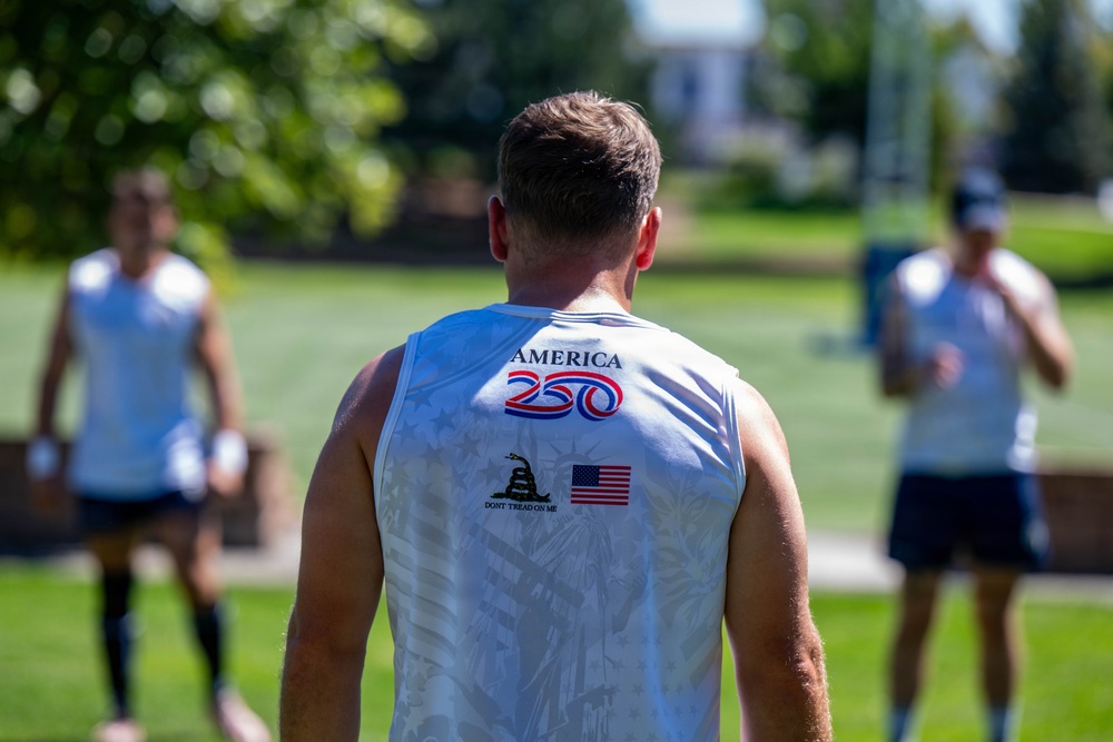 A man is photographed from behind wearing a white sleeveless jersey that reads 'America 250' with a Gadsden flag ('Don't Tread on Me') emblem and a US flag patch. Two other individuals in similar white jerseys are visible in the blurred background on a green grassy field. The setting appears to be an outdoor sports practice or event.