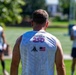 A man is photographed from behind wearing a white sleeveless jersey that reads 'America 250' with a Gadsden flag ('Don't Tread on Me') emblem and a US flag patch. Two other individuals in similar white jerseys are visible in the blurred background on a green grassy field. The setting appears to be an outdoor sports practice or event.