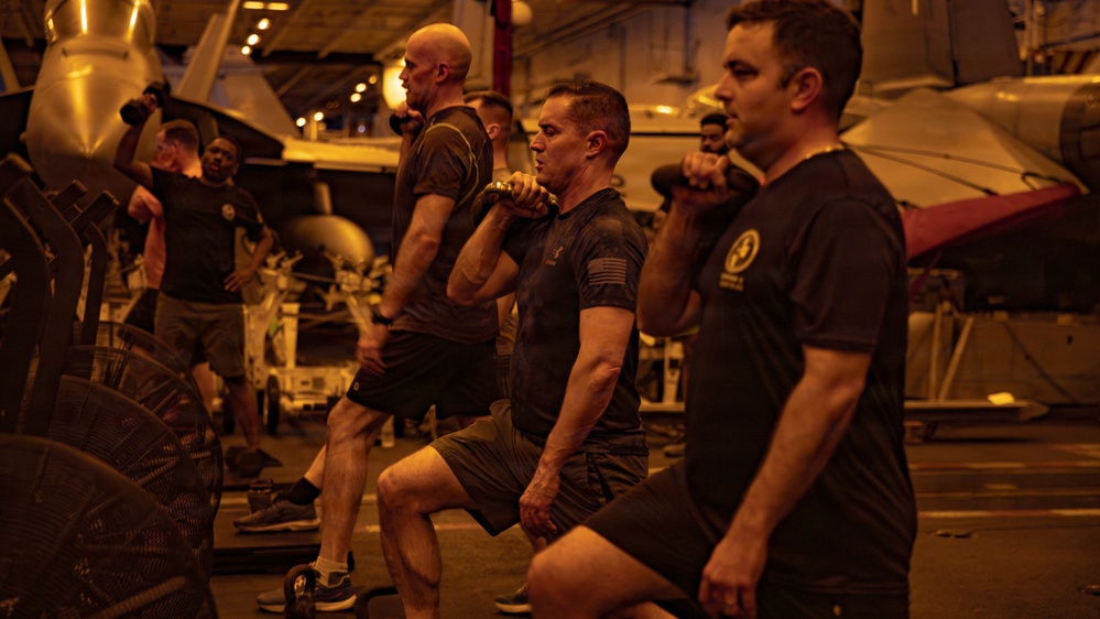 A group of military personnel are performing a synchronized workout exercise, specifically weighted lunges with kettlebells, in what appears to be the hangar bay of a naval aircraft carrier. The space is dimly lit with warm amber lighting and contains visible aircraft and equipment in the background. The participants are wearing matching dark military physical training (PT) uniforms.