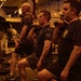 A group of military personnel are performing a synchronized workout exercise, specifically weighted lunges with kettlebells, in what appears to be the hangar bay of a naval aircraft carrier. The space is dimly lit with warm amber lighting and contains visible aircraft and equipment in the background. The participants are wearing matching dark military physical training (PT) uniforms.