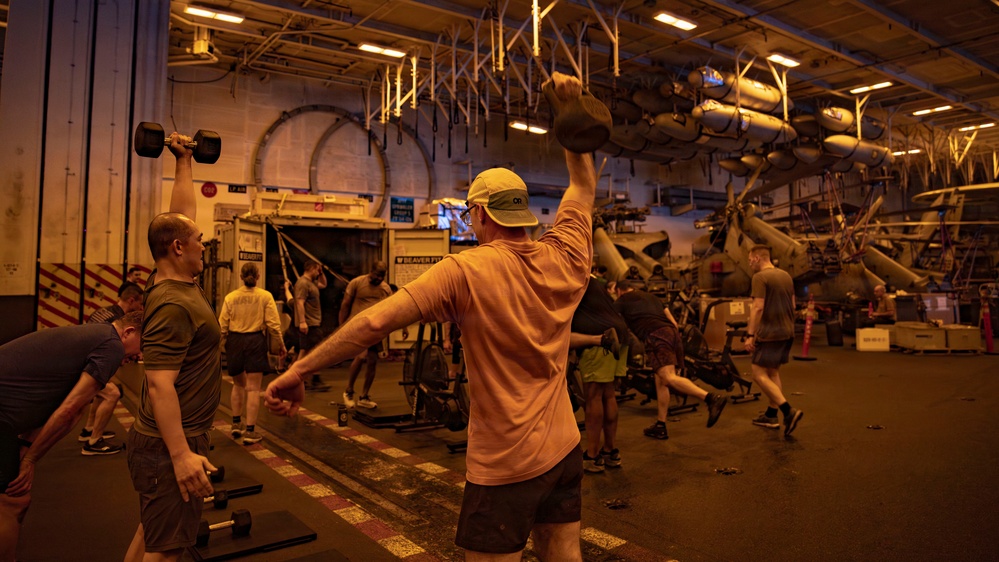 A group of military personnel are conducting a fitness workout session inside what appears to be a naval vessel's hangar bay or flight deck interior. Multiple individuals are performing various exercises including kettlebell presses and dumbbell lifts. Military helicopters and ordnance/equipment are visible in the background, confirming the military ship setting.