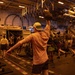 A group of military personnel are conducting a fitness workout session inside what appears to be a naval vessel's hangar bay or flight deck interior. Multiple individuals are performing various exercises including kettlebell presses and dumbbell lifts. Military helicopters and ordnance/equipment are visible in the background, confirming the military ship setting.