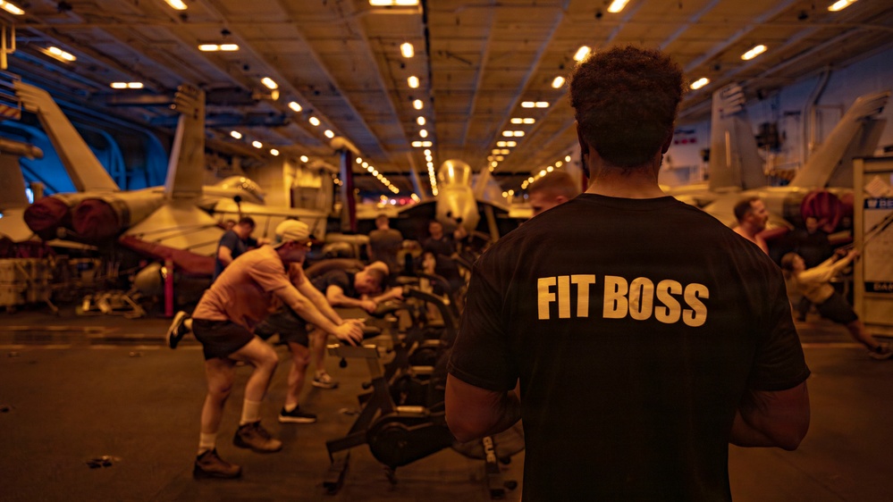 A group of people are exercising in what appears to be the hangar bay of a naval aircraft carrier, surrounded by parked military aircraft. A person in the foreground wearing a black t-shirt with 'FIT BOSS' printed in gold letters on the back appears to be overseeing the workout session. The large enclosed space is dimly lit with overhead lighting, and exercise equipment is visible among the aircraft.