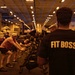 A group of people are exercising in what appears to be the hangar bay of a naval aircraft carrier, surrounded by parked military aircraft. A person in the foreground wearing a black t-shirt with 'FIT BOSS' printed in gold letters on the back appears to be overseeing the workout session. The large enclosed space is dimly lit with overhead lighting, and exercise equipment is visible among the aircraft.