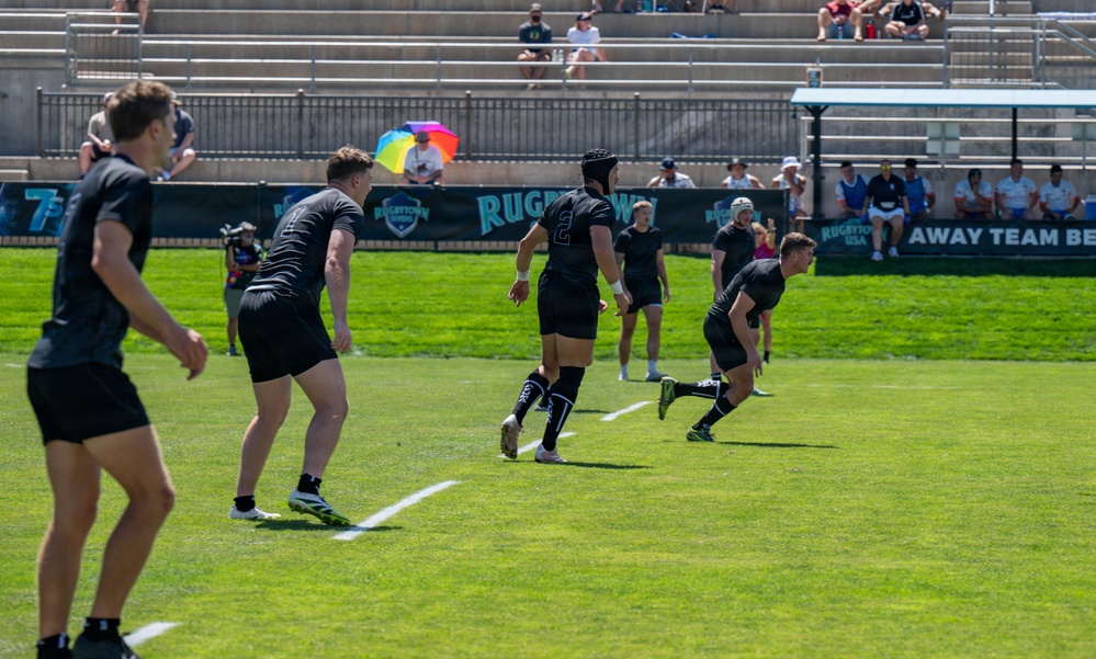 A rugby sevens match is being played on a well-maintained grass field at what appears to be a Rugbytown USA event. Multiple players dressed in all-black uniforms are visible in motion on the field, with spectators watching from stadium bleachers in the background. The game appears to be in active play with players running in formation.