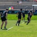 A rugby sevens match is being played on a well-maintained grass field at what appears to be a Rugbytown USA event. Multiple players dressed in all-black uniforms are visible in motion on the field, with spectators watching from stadium bleachers in the background. The game appears to be in active play with players running in formation.
