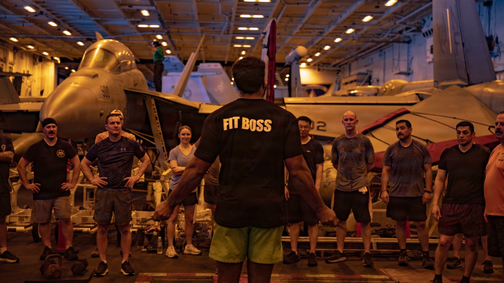 A fitness instructor wearing a black 'FIT BOSS' shirt leads a workout session in the hangar bay of a naval aircraft carrier. A group of approximately 10-12 personnel in athletic wear are gathered in a semicircle facing the instructor. Military jet aircraft, including what appears to be F/A-18 Hornets, are visible in the background.