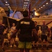 A fitness instructor wearing a black 'FIT BOSS' shirt leads a workout session in the hangar bay of a naval aircraft carrier. A group of approximately 10-12 personnel in athletic wear are gathered in a semicircle facing the instructor. Military jet aircraft, including what appears to be F/A-18 Hornets, are visible in the background.
