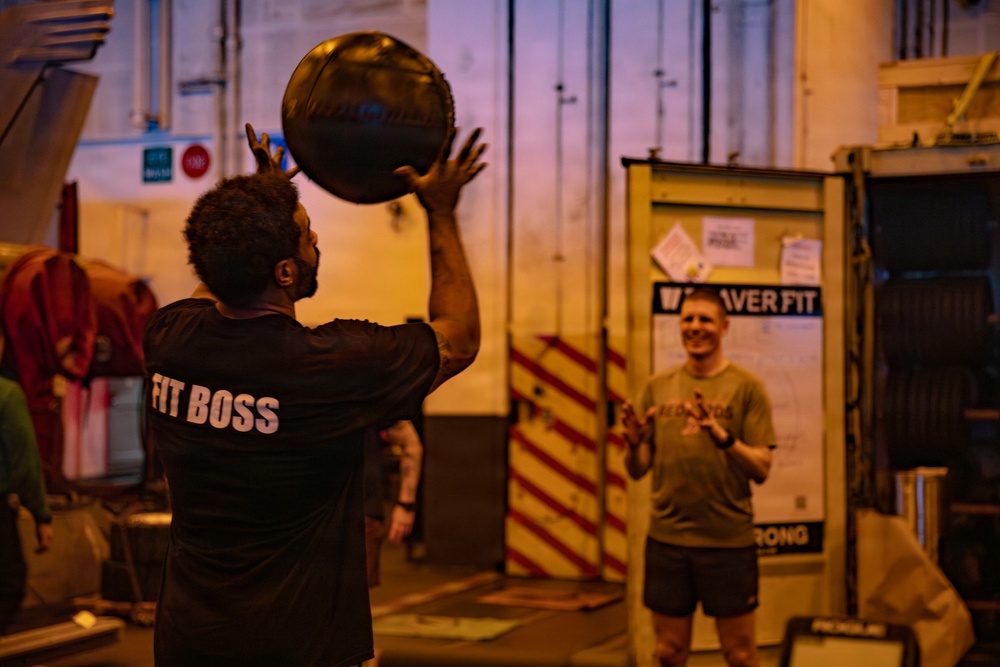 Two individuals are engaged in a medicine ball exercise in what appears to be an industrial or warehouse-style gym space. The person in the foreground is wearing a black shirt reading 'FIT BOSS' and is tossing or catching a dark medicine ball overhead, while a second person in the background smiles and appears ready to receive or has just thrown the ball. The setting has warm amber lighting and features safety-striped pillars and signage including 'WAIVER FIT' and 'STRONG'.