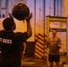 Two individuals are engaged in a medicine ball exercise in what appears to be an industrial or warehouse-style gym space. The person in the foreground is wearing a black shirt reading 'FIT BOSS' and is tossing or catching a dark medicine ball overhead, while a second person in the background smiles and appears ready to receive or has just thrown the ball. The setting has warm amber lighting and features safety-striped pillars and signage including 'WAIVER FIT' and 'STRONG'.