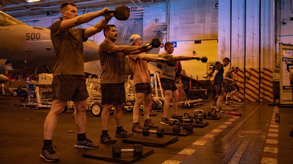 A group of military personnel are conducting a fitness workout session in what appears to be the hangar bay of an aircraft carrier. Multiple individuals are performing kettlebell swings in a synchronized line, with military aircraft and equipment visible in the background. The space has been temporarily converted into a workout area using rubber mats and various free weights.