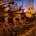 A group of military personnel are conducting a fitness workout session in what appears to be the hangar bay of an aircraft carrier. Multiple individuals are performing kettlebell swings in a synchronized line, with military aircraft and equipment visible in the background. The space has been temporarily converted into a workout area using rubber mats and various free weights.