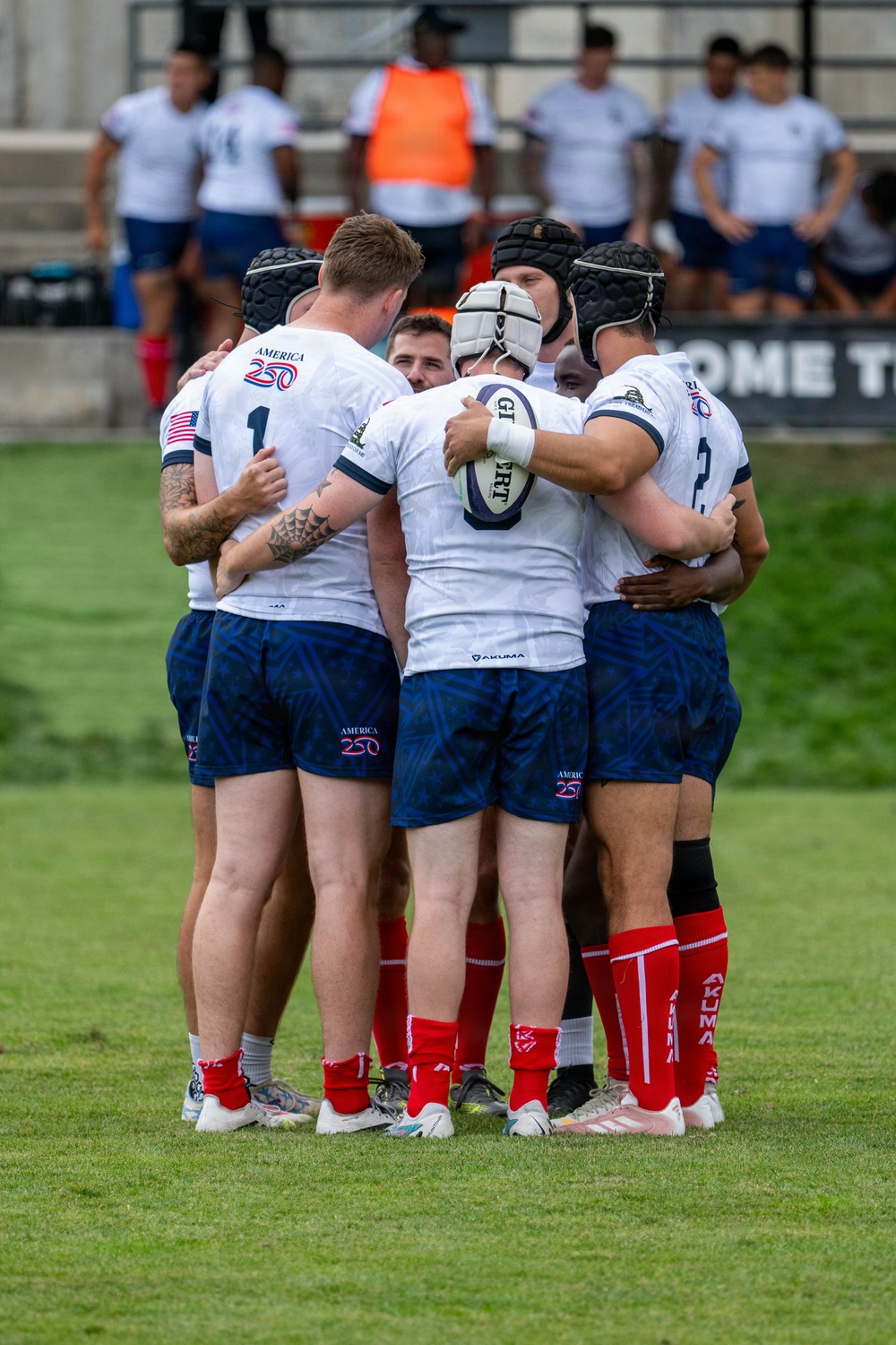 A rugby team huddles together on a grass field during a match or training session. The players are wearing white jerseys with 'America 250' branding, blue patterned shorts, red socks, and several players wear protective headgear. One player in the center of the huddle is holding a rugby ball.