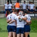 A rugby team huddles together on a grass field during a match or training session. The players are wearing white jerseys with 'America 250' branding, blue patterned shorts, red socks, and several players wear protective headgear. One player in the center of the huddle is holding a rugby ball.