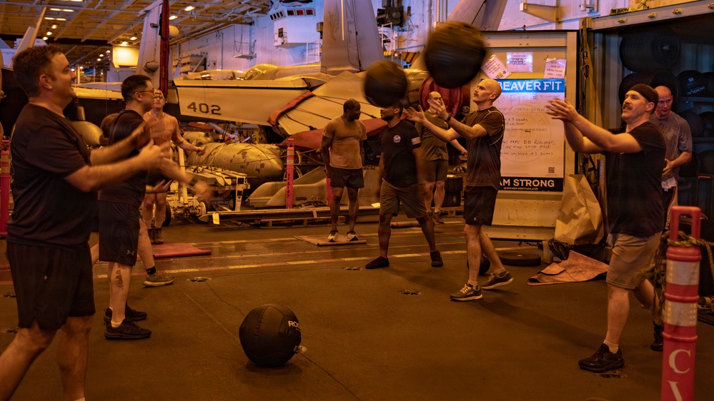 Military personnel are conducting a fitness workout session in the hangar bay of a naval aircraft carrier. Sailors are performing medicine ball exercises in a group setting, with a parked military aircraft (numbered 402) and fitness equipment visible in the background. A 'Beaver Fit' branded fitness station and a whiteboard with workout instructions are visible to the right.